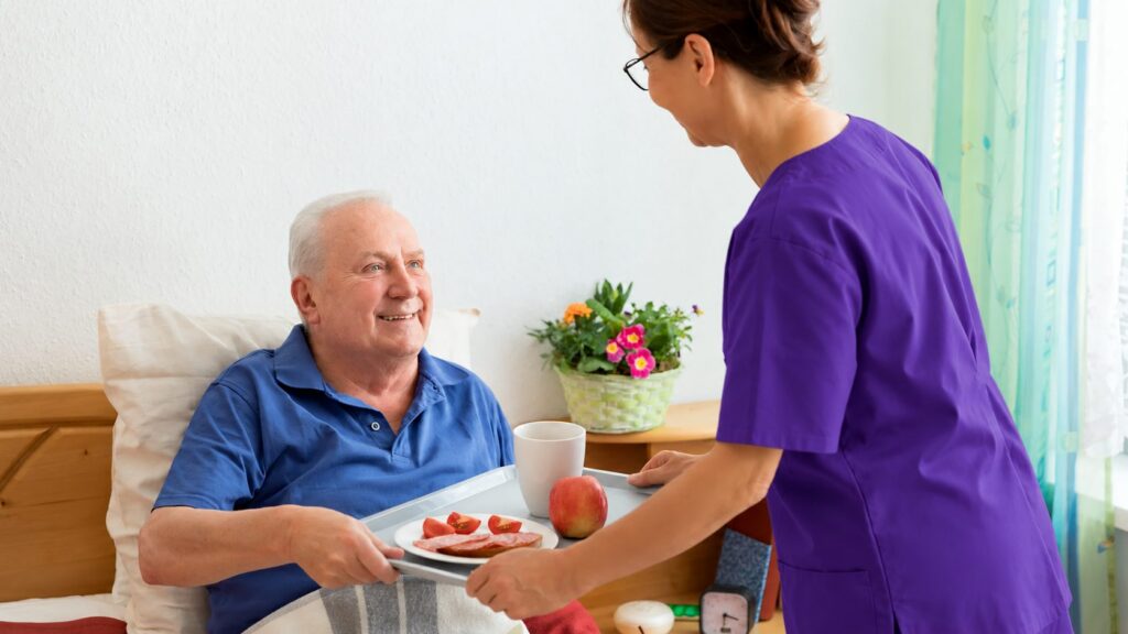 Caregiver in purple scrubs bringing a meal tray to a senior man resting in bed